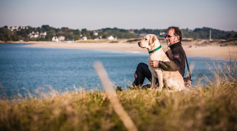 man and yellow labrador sitting on grass next to the beach